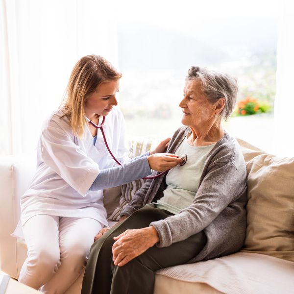 nurse sits next to woman on couch and listens to heart with stethoscope